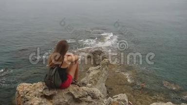 女游客背着背包坐在晴朗的天空和平静的海洋背景上的岩石上